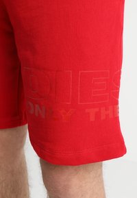 Shorts rouges arrivant aux genoux avec un texte discret en relief sur le côté, portés par une personne à la peau claire et aux poils de jambes.