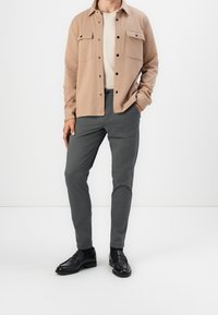Uomo che indossa una camicia beige sbottonata sopra una t-shirt crema, pantaloni slim grigio scuro e mocassini in pelle nera, in piedi con una mano in tasca.