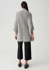 Poncho tissé en motif pied-de-poule noir et blanc, doté d'un col haut, de manches larges et d'une coupe décontractée. Associé à un pantalon noir et des chaussures.
