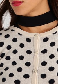 Cárdigan beige con lunares negros, con un frente abotonado y un escote de gargantilla negro. Tejido suave con una textura lisa.