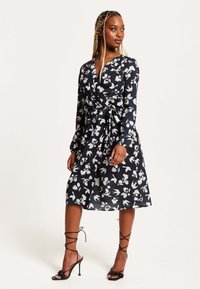 Wrap dress in navy fabric with white floral pattern, long sleeves, and a tie at the waist. Paired with strappy black heels.
