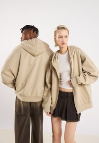Beige overdimensjonerte hettegensere med glidelås foran. Den mannlige modellen har på seg mørke, vide bukser, mens den kvinnelige modellen har på seg et svart plisségjort mini-skjørt.