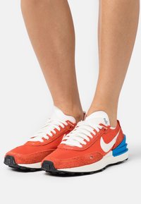 Red textile sneakers with white laces and swoosh logo. Features a blue heel accent and black textured sole. Suede and mesh materials.