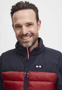 Veste matelassée bleu marine et rouge avec col montant, fermeture éclair à l'avant et écusson logo. Tissu texturé avec une finition lisse en haut et un bas matelassé.