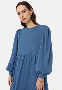 Blue pleated dress with long puff sleeves, round neckline, and a flowing silhouette. Fabric has a soft, textured finish.