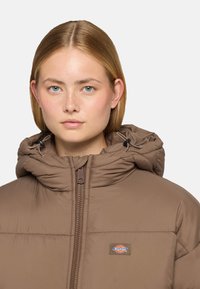 Chaqueta acolchada marrón con capucha y cierre de cremallera, que presenta un parche de logo. La tela tiene un aspecto texturizado y la capucha está fruncida con cordones.