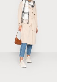 Kobieta w beżowym trenczu, niebieskich, krótkich dżinsach, białych sneakersach, w kratkowanym szaliku, trzymająca małą brązową torebkę na tle białym.