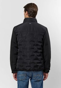 Mann iført en svart quiltet jakke med høy krage og blå jeans, vist bakfra mot en enkel lys bakgrunn.