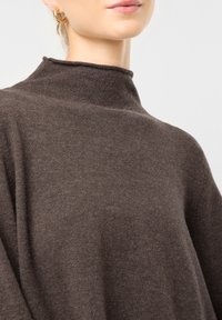 Hnědý svetr s vysokým límcem, vyrobený z měkké tkaniny s uvolněným střihem. Detailní záběr zdůrazňuje vlastnosti látky a hladký, rovnoměrný povrch.