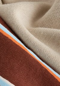 Megztas audinys, kuriame dera smėlio spalvos skyrius su plataus rudo ruožo ir oranžinių bei šviesiai mėlynų juostelių intarpais, demonstruojantis minkštą, tekstūruotą paviršių.