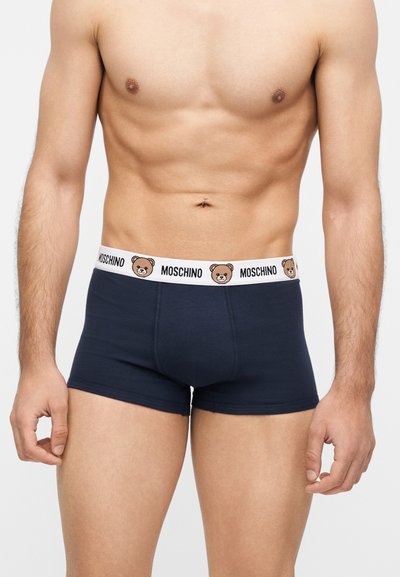 Αρσενικός κορμός που φοράει σκούρα μπλε μπλέιζερ με λευκή μέση με κείμενο Moschino και πρόσωπα αρκουδάκια.