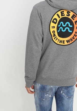 Personne portant un sweat à capuche gris avec un logo rond orange et noir affichant "Diesel Only The Waves" et un jean bleu déchiré.