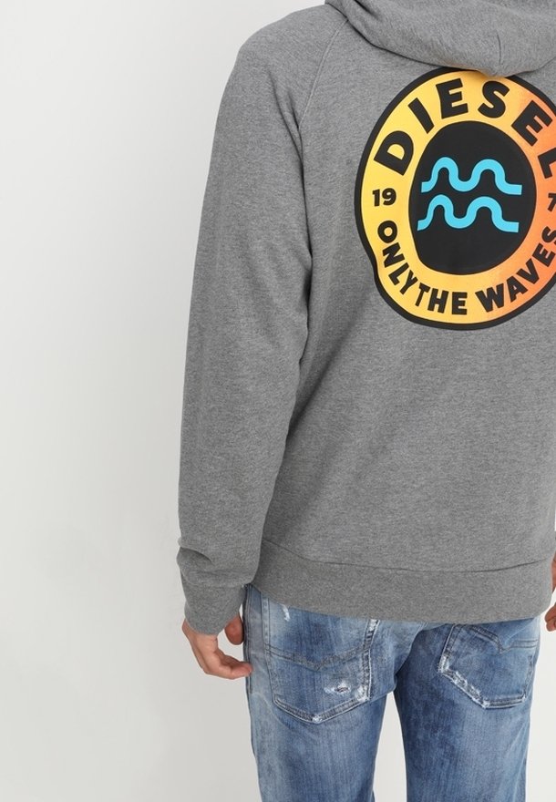 Personne portant un sweat à capuche gris avec un logo rond orange et noir affichant "Diesel Only The Waves" et un jean bleu déchiré.