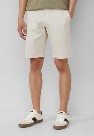 Light beige cotton shorts with a straight cut, side pockets, and rolled hems, paired with white and brown athletic sneakers.