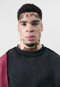 Schwarz und burgunderfarbener Sweatshirt mit einem ausgefransten Ausschnitt und Akzenten, aus einem strukturierten Stoff gefertigt und mit einem kleinen Logo auf der Brust.