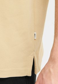 Beige teksturert polo-skjorte med sidesplitt og en hvit etikett med utropstegn. Utsikten fokuserer på den nedre siden og hånddetaljen.