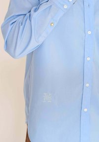 Chemise bleu clair à boutons, fabriquée en tissu lisse, avec des boutons blancs et un détail brodé subtil sur le côté gauche.