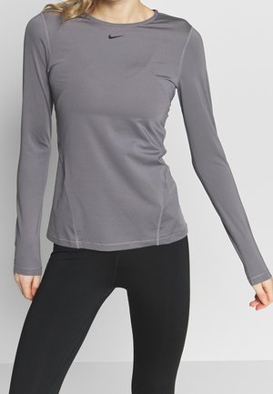 Frau trägt ein eng anliegendes graues, langärmliges Sportshirt mit schwarzem Nike-Logo und schwarze Leggings, steht vor einem einfarbigen Hintergrund.