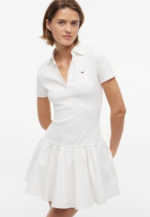 Kobieta o krótkich brązowych włosach, ubrana w biały, krótki rękaw, sukienkę z kołnierzykiem i marszczonym dołem, stojąca na jednolitym białym tle.