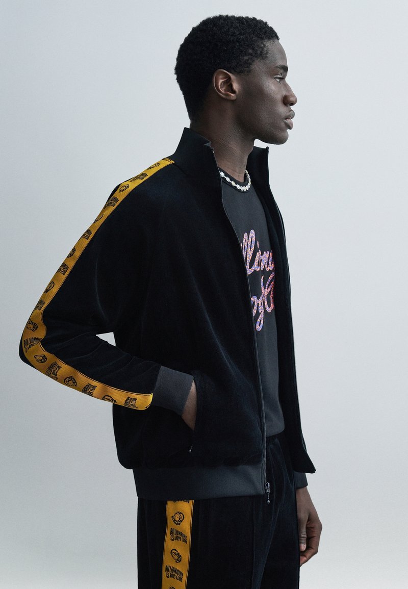 Μαύρο βελούδινο τζάκετ φόρμας με κ ίτρινη ταινία λογότυπου κατά μήκος των μανικιών, κλείσιμο με φερμουάρ και ριμπ μανσέτες. Συνδυάζεται με μαύρο t-shirt με γυαλιστερά γράμματα.