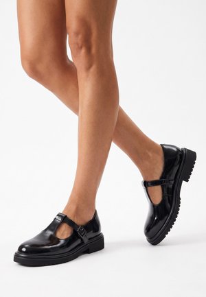 Piernas con zapatos Mary Jane negros brillantes con correas de hebilla y suelas de goma gruesas sobre un fondo blanco.