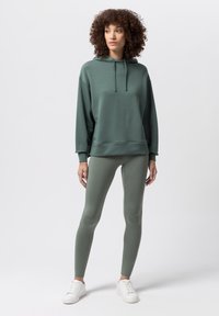 Zielona bluza z kapturem, z sznurkami, o luźnym kroju i ściągaczu, w zestawieniu z dopasowanymi legginsami i białymi trampkami.