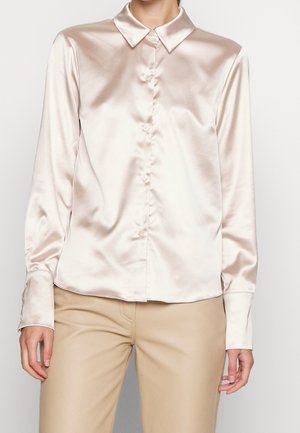 Femme portant une chemise en satin crème à manches longues boutonnée avec un pantalon beige, visible de la poitrine aux hanches sur un fond uni.
