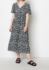 Svartvit klänning med bladmotiv, midi-längd, korta ärmar, V-ringning, knappfront och luftig silhuett, tillsammans med svarta sandaler.