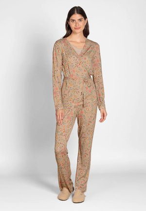 Woman standing wearing beige floral long-sleeve pajama set with wrap top and matching pants, beige slippers, against plain light background.