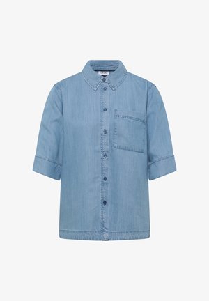 Kvinde iført løs, lyseblå denimskjorte med albuelange ærmer, set bagfra med synlige detaljer ved skulderstykke og central læg.