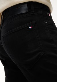 Κοντινό πλάνο μαύρου τζιν Tommy Hilfiger με λογότυπα στο πίσω τσεπάκι και τη μέση, φορεμένο με μπεζ τοπ.