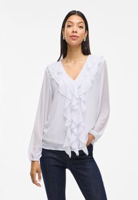 Hvit chiffon-bluse med V-hals og en fremtredende volangdetalj foran. Lange ermer med rynkede mansjetter, kombinert med mørke jeans.