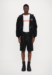Black hooded sweatshirt with white stitching, graphic white shirt, black shorts with contrast stitching, and knee-high black boots.