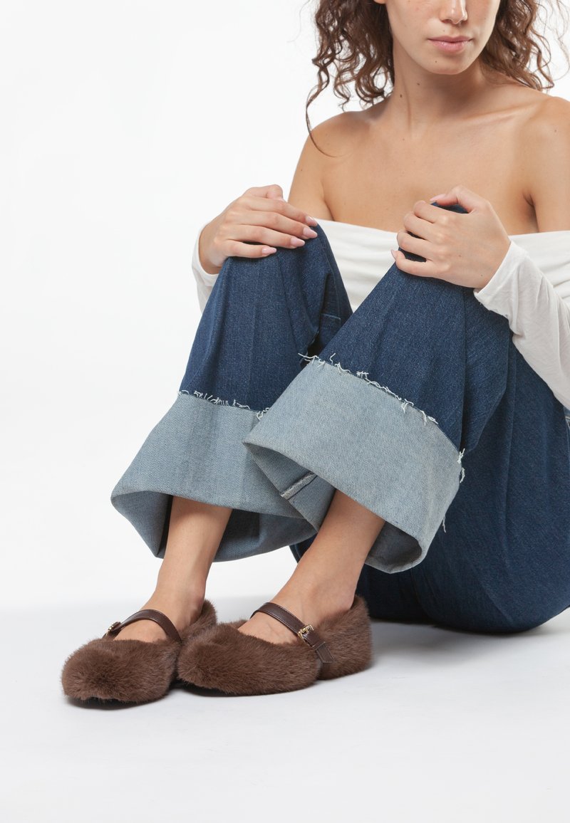 Pantofole marroni pelose con punta arrotondata, dotate di una fascia marrone e una fibbia. Jeans scuri larghi con polsini in denim chiaro e orlo sfilacciato.