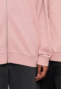 Δύο άτομα φορούν ανοιχτό ροζ φούτερ με φερμουάρ μπροστά και τσέπες, καθώς και σκούρα γκρι τζιν, εμφανιζόμενα από τη μέση έως το χέρι.