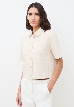 Vrouw met schouderlang donker haar, gekleed in een gestructureerd beige kortemouwig overhemd met knopen en een witte broek, met één hand in de zak.
