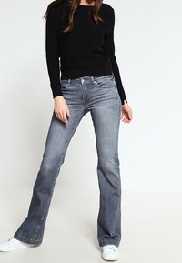 Kobieta w czarnym długim rękawie, szarych dżinsach bootcut i białych trampkach, stojąca z nogami rozstawionymi na tle białego tła.