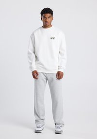 Felpa bianca con scollatura a girocollo e una piccola toppa grafica colorata. Abbinata a pantaloni della tuta grigi chiari e sneakers bianche.