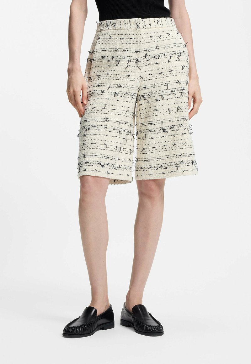 Shorts crème avec un motif parsemé noir, en tissu tissé texturé, taille élastique et coupe mi-genou. Chaussures noires à enfiler.