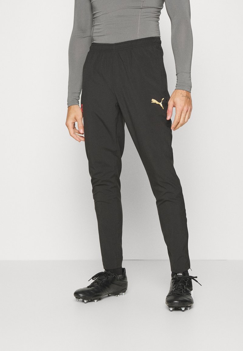 Pantalon de sport noir avec taille élastique, forme fuselée, accent du logo doré de Puma et texture lisse. Associé à des crampons noirs.