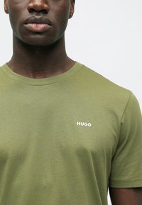 Olivgrünes T-Shirt mit Rundhalsausschnitt aus weichem Stoff, mit kleinem weißem "HUGO"-Logo auf der linken Brust. Glatte Textur, kurze Ärmel.