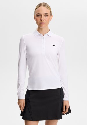 Donna che indossa una polo bianca a maniche lunghe e una gonna nera al ginocchio, rivolta verso di noi su uno sfondo chiaro e uniforme.