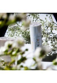 Vitt cylindriskt hudvårdsprodukt med guldtext, detaljerat mönster på locket, omgiven av små vita blommor och en reflekterande yta.