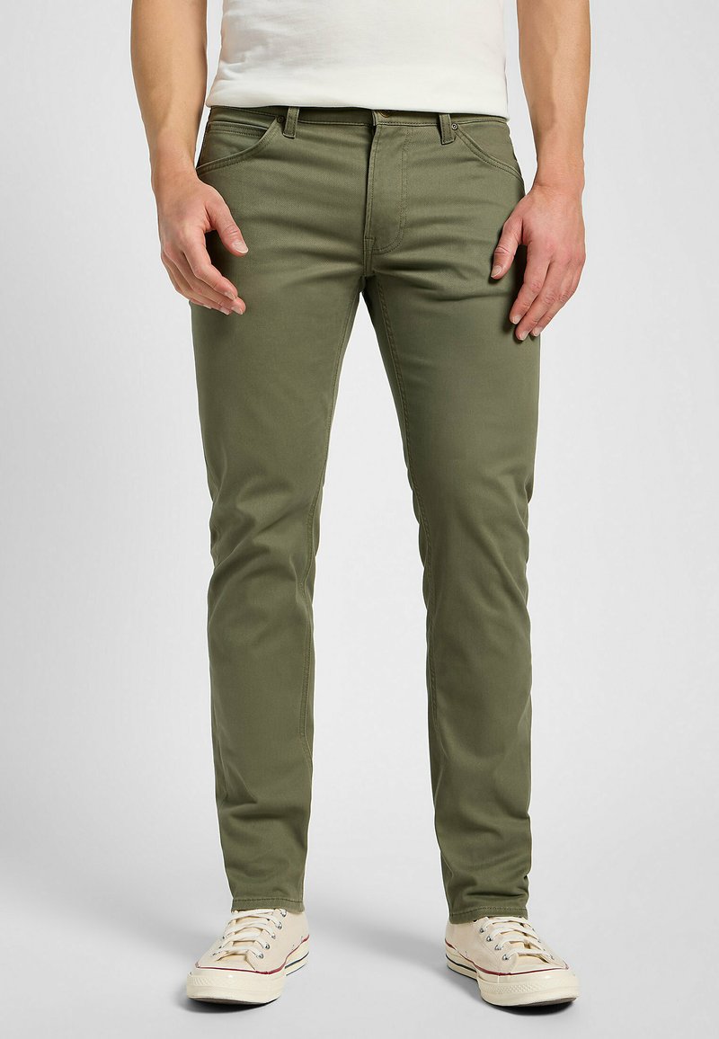 Pantalones de algodón elástico verde oliva con cinco bolsillos, diseño ajustado, costuras sutiles, y combinados con zapatillas de lona beige.
