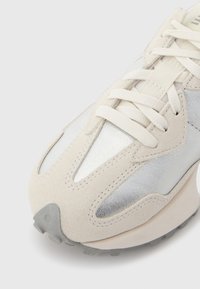 Κοντινό πλάνο στη μύτη ενός αθλητικού παπουτσιού σε μπεζ και ασημί χρώμα, με λευκά κορδόνια και υφασμάτινες σουέτ λεπτομέρειες, σε λευκό φόντο.