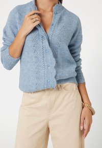 Vrouw in een lichtblauwe gebreide cardigan met knopen en beige broek, waarbij ze haar hand toont met een ring en een gouden armband.