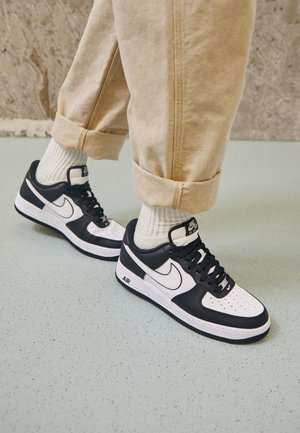 Osoba w beżowych podwiniętych spodniach, białych prążkowanych skarpetkach i czarno-białych sneakersach Nike Air Force 1 stoi na jasnoszarej podłodze.