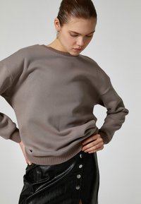 Brązowy sweter dzianinowy z okrągłym dekoltem i ściągaczami, zestawiony z czarną spódnicą z ekologicznej skóry z zatrzaskami.