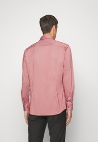 Uomo vestito con una camicia rosa a maniche lunghe e bottoni e pantaloni neri, in piedi con la schiena rivolta verso uno sfondo bianco semplice.