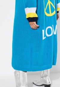 Turkoosi neule oversized villatakki, jossa on "LOVE" valkoisilla kirjaimilla ja rauhanmerkki keltaisena, sekä moniväriset raidoitetut hihat.
